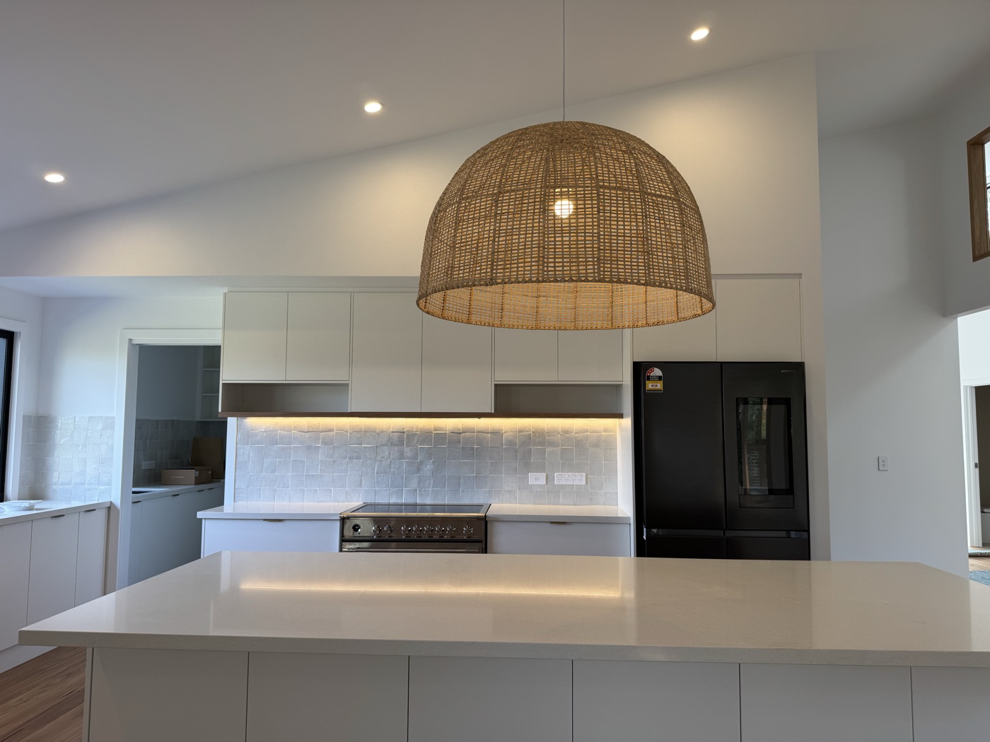 A finished kitchen with a rattan dome pendant over the bench, downlights across the ceiling, and warm strip lighting under the overhead cabinets